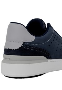Sneaker in suede blu navy con un tallone grigio a contrasto, design perforato, suola bianca e cuciture a contrasto. Logo Clarks visibile sul tallone.