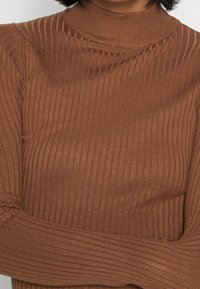 Kobieta w brązowym swetrze z długim rękawem i golfem, z założonymi rękami, prezentująca fakturę materiału i wzór dzianiny.