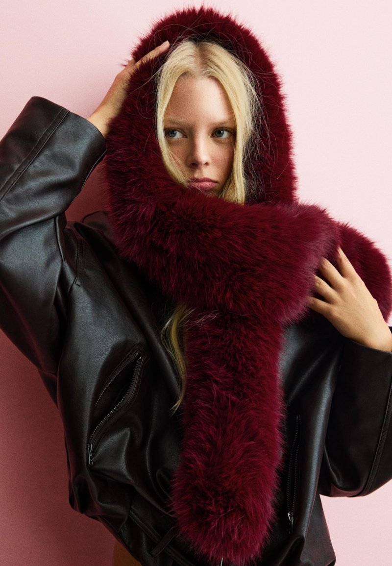 Bordo umjetni krzneni šal prebačen preko modela koji nosi crnu kožnu jaknu, postavljen na ružičastoj pozadini. Mekoća i bujan oblik.