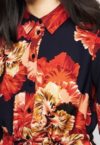 Blusa floreale con grandi fiori rossi e arancioni vivaci su uno sfondo blu scuro. Include un colletto e dettagli con bottoni.
