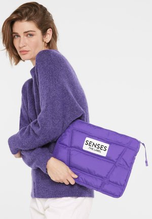 Lila, gesteppt Laptop-Hülle mit Reißverschluss, die ein weißes Etikett mit der Aufschrift "SENSES - THE LABEL" zeigt. Der Benutzer hält sie gegen einen gemütlichen lila Pullover.