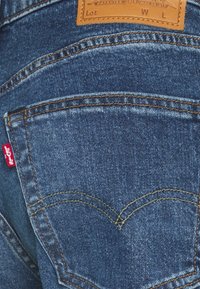 Jeans de mezclilla azul oscura con costuras amarillas en contraste, bolsillos traseros clásicos y una etiqueta roja de Levi's cerca del bolsillo izquierdo.