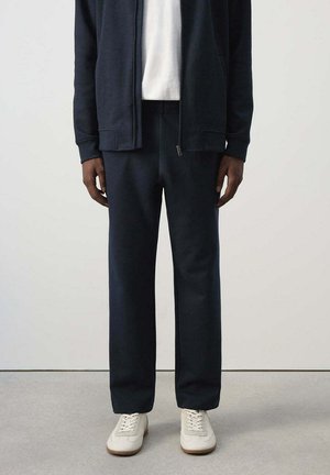 Personne portant un pantalon et une veste bleu marine avec des baskets blanches, debout contre un mur et un sol clairs et unis.