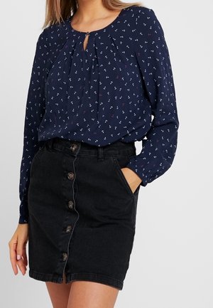 Woman wearing a navy long-sleeve blouse with small anchor prints tucked into a high-waisted black button-front denim skirt.
