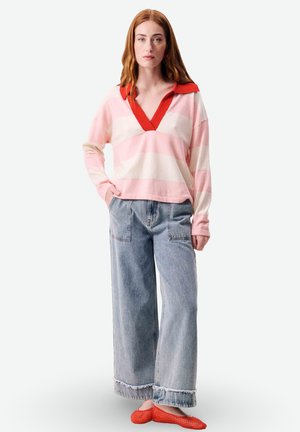 Jeune femme aux longs cheveux roux portant un pull rayé rose et blanc avec col rouge, un jean à jambes larges et des ballerines rouges, debout les mains dans les poches.