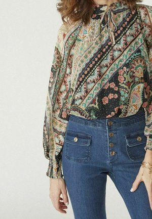 Femme portant un chemisier à manches longues à motif floral avec un col à nouer et un jean bleu taille haute avec des détails de boutons et de poches.