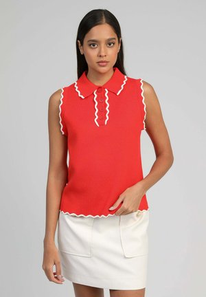 Femme portant un polo rouge sans manches avec un bord ondulé blanc, associé à une jupe blanche, debout devant un fond gris clair.