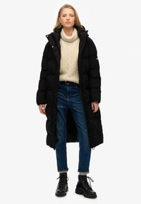 Abrigo largo negro de plumas con capucha, diseño acolchado, botones a presión, llevado sobre un suéter de punto en color crema y jeans azules, combinado con botas negras con cordones.