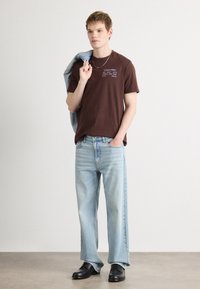 Camiseta marrón de manga corta con texto blanco, jeans rectos de mezclilla azul claro y zapatos negros, con una chaqueta de mezclilla colgando de un hombro.