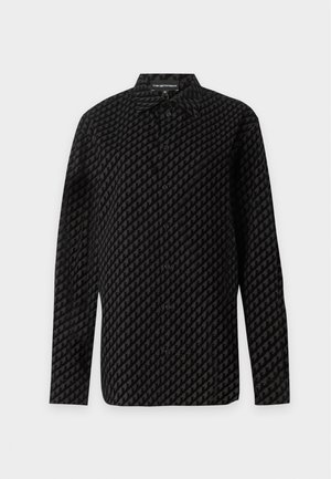 Femme debout tenant une pochette noire, portant une chemise noire à motifs oversize, un jean bleu à jambes larges et des ballerines noires pointues sur fond uni.