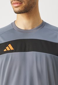 Sportovní tričko s šedým tělem, černým horizontálním pruhem a oranžovými logo akcenty. Hladký materiál s kulatým výstřihem a krátkými rukávy.