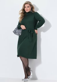Abito midi verde scuro, con maniche lunghe, collo alto e cintura regolabile. Indossato con scarpe argentate con tacco alto e una pochette di paillettes.
