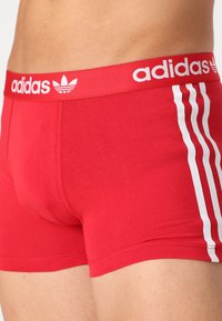Rote Baumwoll-Boxershorts mit weißem Adidas-Logo-Bund und drei vertikalen weißen Streifen an der Seite. Weicher Stoff, figurbetontes Design.