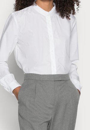 Kobieta w białej koszuli zapinanej na guziki z falbankowym kołnierzykiem i szarych spodniach z wysokim stanem, ręce w kieszeniach.