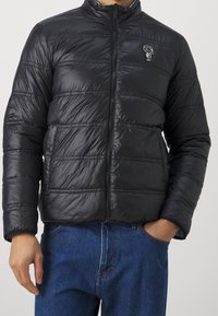 Svart puffjacka med hög krage, horisontell sömnad, dragkedjestängning och en liten grafisk accent på bröstet. Buren över blå jeans.