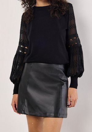 Femme portant un haut noir avec des manches bouffantes transparentes en dentelle et une mini-jupe patineuse en cuir noir, posant devant un fond neutre.