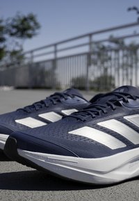 Scarpe da corsa blu navy con una tomaia testurizzata, design a tre strisce bianche e suola leggera. Inquadratura ravvicinata sulle stringhe e sui dettagli della soletta.