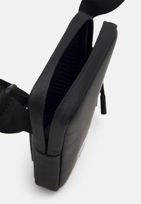 Borsa in tessuto nero con superficie testurizzata, forma rettangolare piatta, chiusura con zip e tracolla regolabile. Interno foderato in materiale a rete.