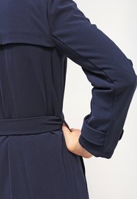 Abrigo azul marino de tela suave, con cintura ceñida, mangas largas y detalle de yugo en la parte posterior.