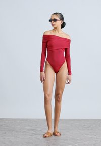 Punane õlgadeta pika varrukaga ujumistrikoo kõrge lõike ja siledapinnalise ning minimalistliku disainiga, kombineeritud pruuni tasapinnaliste sandaalidega.