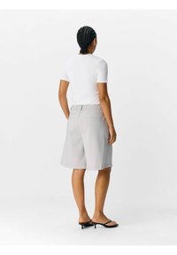 Pantaloni corti grigi su misura con vestibilità relax, dotati di vita elasticizzata e tasche laterali. Abbinati a una camicia bianca attillata e sandali neri.