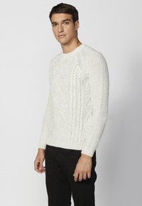 Weißer Strickpullover mit einem Zopfmuster, rundem Halsausschnitt und gerippten Bündchen. Kombiniert mit schwarzen Hosen, getragen von einer stehenden Person.