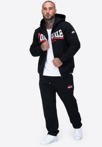 Sudadera con cremallera negra con un gran logotipo en blanco y rojo, combinada con pantalones de chándal negros que presentan un pequeño logotipo y zapatillas blancas. Textura de tela suave.