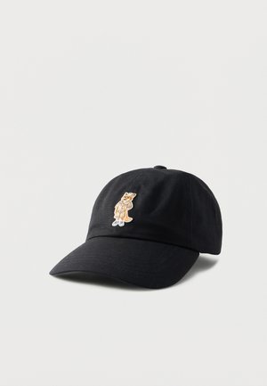 Gorra de béisbol negra con visera curva que presenta un pequeño zorro bordado que lleva un abrigo beige y zapatos blancos en el centro delantero.
