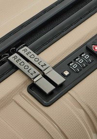 Primer plano de una maleta texturizada beige con una cerradura de combinación TSA negra y dos tiradores de cremallera plateados grabados con "REDOLZ".