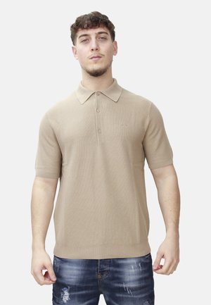 Giovane uomo con capelli corti e ricci che indossa una polo beige a maniche corte e jeans blu usati, in piedi davanti a uno sfondo bianco.