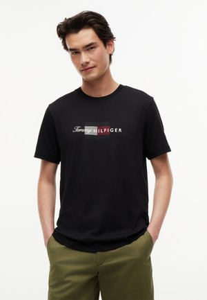 Junger Mann trägt ein schwarzes Tommy Hilfiger Logo-T-Shirt und olivgrüne Hose, steht mit den Händen in den Taschen vor weißem Hintergrund.