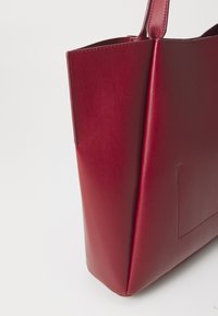 Rød skinn totebag med et stilrent, strukturert design. Har glatt tekstur, minimalisk stil og et enkelt, solid håndtak.