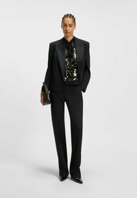 Schwarzer maßgeschneiderter Anzug mit weiten Hosen, einem mit Pailletten besetzten Oberteil und einer Fliege. Kombiniert mit spitzen schwarzen High Heels und einer kleinen schwarzen Handtasche.
