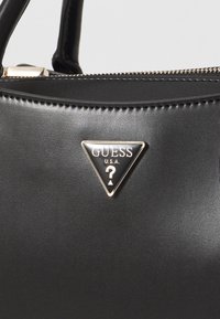 Mustapintainen synteettinen käsilaukku, jossa on kolmion muotoinen Guess-logo metallisilla yksityiskohdilla. Tasainen pinta, jossa on vetoketjukiinnitys ja kontrastiväriset ommelkoristeet.