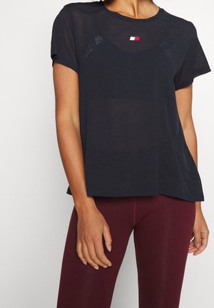 Kobieta ubrana w czarną koszulkę z krótkim rękawem z małym logo w kolorach czerwonym, białym i niebieskim, w połączeniu z bordowymi legginsami, stojąca na białym tle.