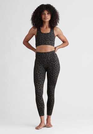Frau mit lockigem Haar steht barfuß und trägt einen passenden dunkel gemusterten Leoparden-Sport-BH und hoch taillierte Leggings vor einem schlichten Hintergrund.