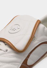 Baskets en cuir blanc et marron avec logo cousu sur la languette rembourrée et panneau latéral perforé, mettant en valeur un savoir-faire détaillé.