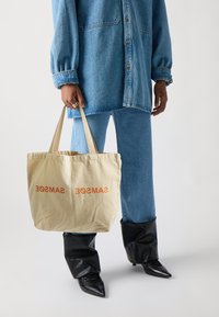 Kanna uz audekla maisiņa, smilškrāsā, ar uzrakstu "SAMSØE" oranžā krāsā, ko nes cilvēks džinsu apģērbā un melnās, norādītās zābakos.