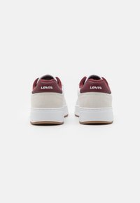 Bijele tenisice s maroon naglasima i bež suknjenim panelima. Imaju debeli bijeli potplat i istaknut Levi's logo na poleđini.