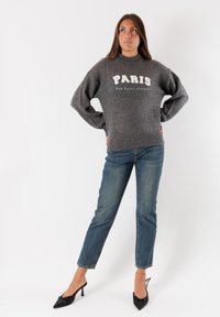 Donna che indossa un maglione grigio "PARIS Rue Saint-Honoré", jeans blu e tacchi neri, in piedi con le mani sui fianchi su uno sfondo bianco.