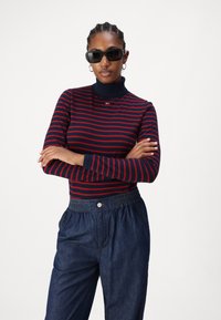 Maglione a collo alto a righe navy e rosse con maniche lunghe, abbinato a pantaloni in denim scuro con vita elastica e dettaglio di bottone frontale.