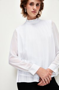 Blusa bianca plissettata con scollo alto, maniche lunghe trasparenti e polsi con bottoni. Texture liscia, design stratificato e vestibilità ampia.