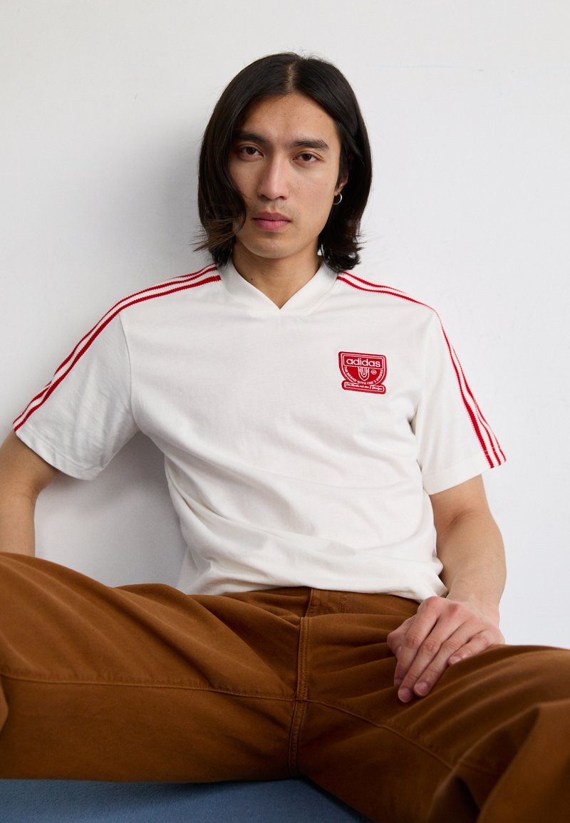 Camisa branca de mangas curtas com detalhes em vermelho e logótipo da adidas, com um design de decote em V. Modelo a usar calças castanhas, sentado numa superfície azul.