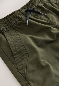 Olivovo zelené šortky vyrobené z mäkkej tkaniny. Majú elastický pás so šnúrkou a bočné vrecká s dekoratívnym prešívaním.