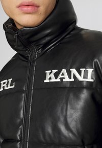 Črna usnjena puffer jakna z visokim ovratnikom, zadrgo in belimi vezenimi logotipi "KARL" in "KANI" spredaj.