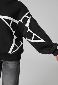 Svart sweatshirt med vitt grafiskt tryck, med rundad hals och ribbade ärmslut. Tyget verkar mjukt med en avslappnad passform.