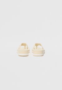 Cremefarvede flip-flops med fletstraps og en flad sål, der har en struktureret fodseng og hvide sålaccenter.