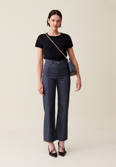 T-shirt noir à col rond, jeans évasés en denim foncé taille haute avec des coutures visibles, associés à des talons aiguille noirs et un petit sac.