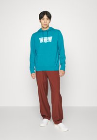 Teal hoodie met een wit logo, voorzak en verstelbare koordjes, gecombineerd met losse, roestkleurige broeken en witte sneakers.