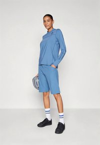 Világoskék sportos öltözet, amely hosszú ujjú felsőből és térdig érő rövidnadrágból áll, nedvességelvezető anyagból készült. Fekete cipő és sisak.